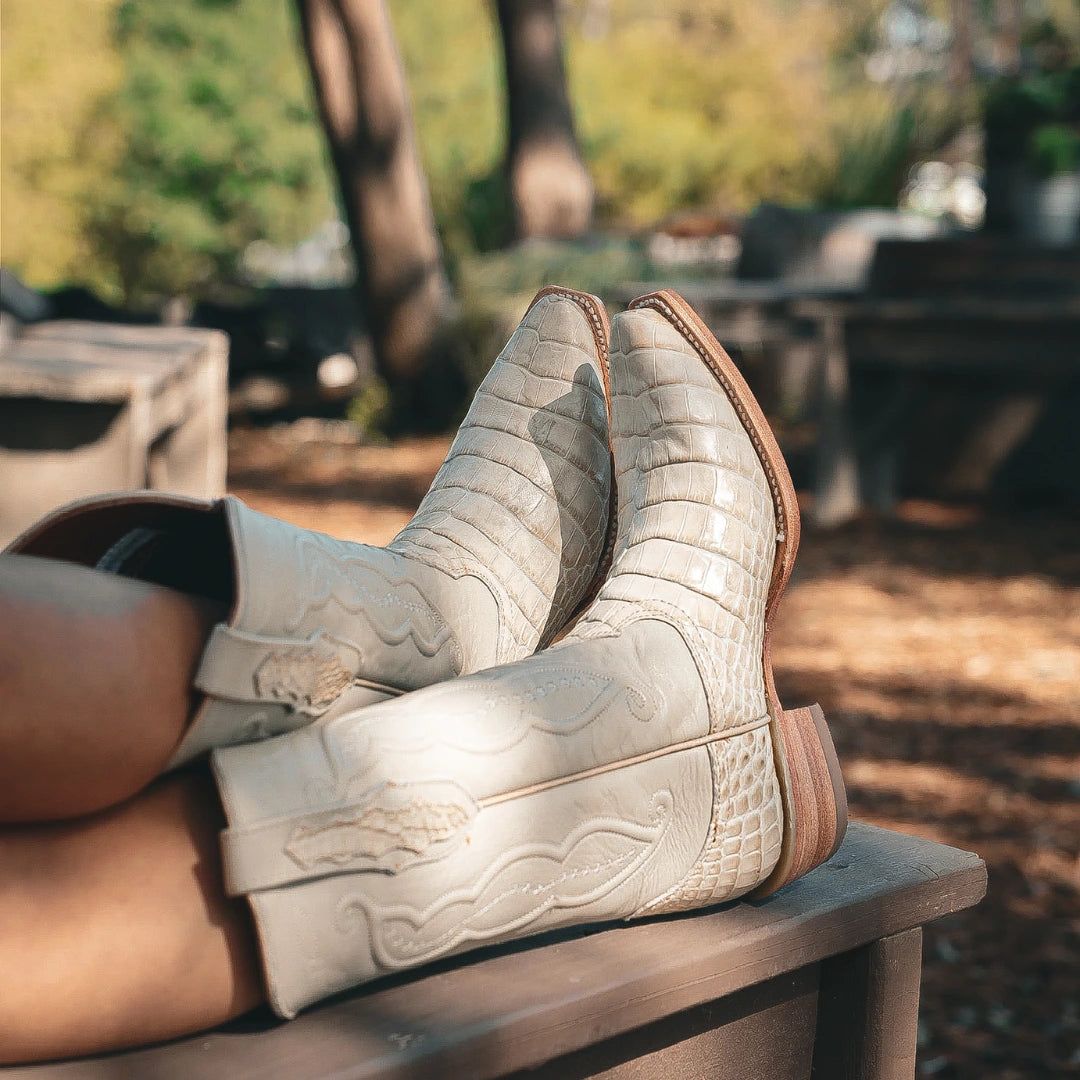 A pair of ivory alligator belly leather cowboy boots with brown soles, worn on a person's feet resting on a wooden bench in an outdoor park setting.