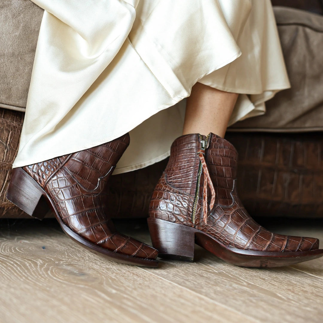 A pair of mocha alligator belly ankle boots with a side zipper and tassel detail, worn with a cream satin skirt on a wooden floor.