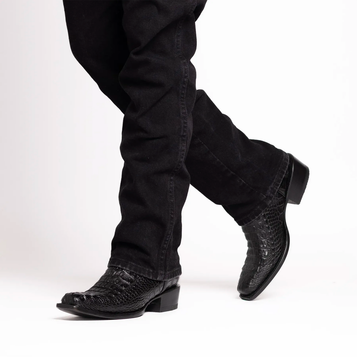 Black alligator cowboy boots worn with black jeans, shown from the calves down against a white studio background.