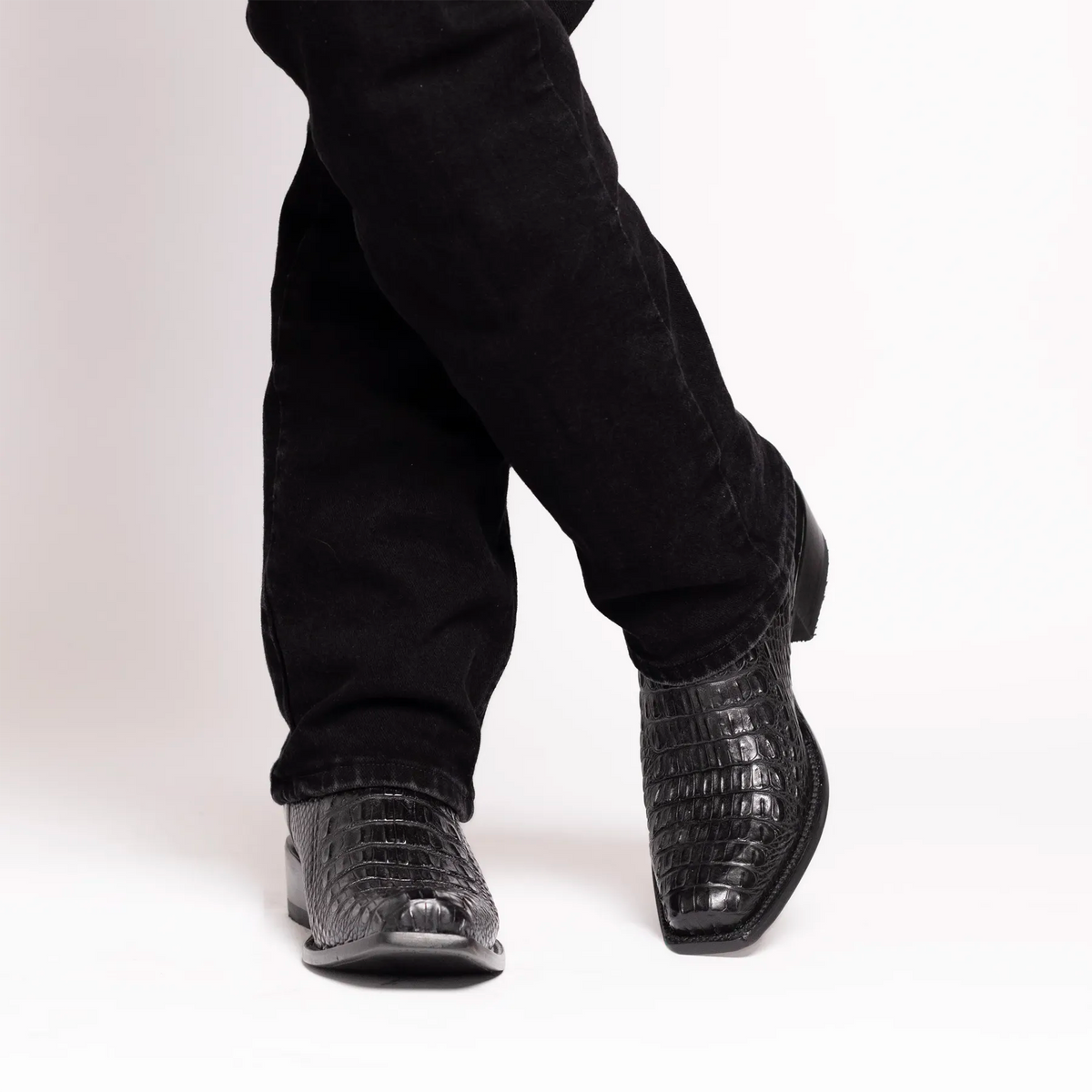 Black alligator cowboy boots worn with black jeans, shown from a side angle against a white studio background.
