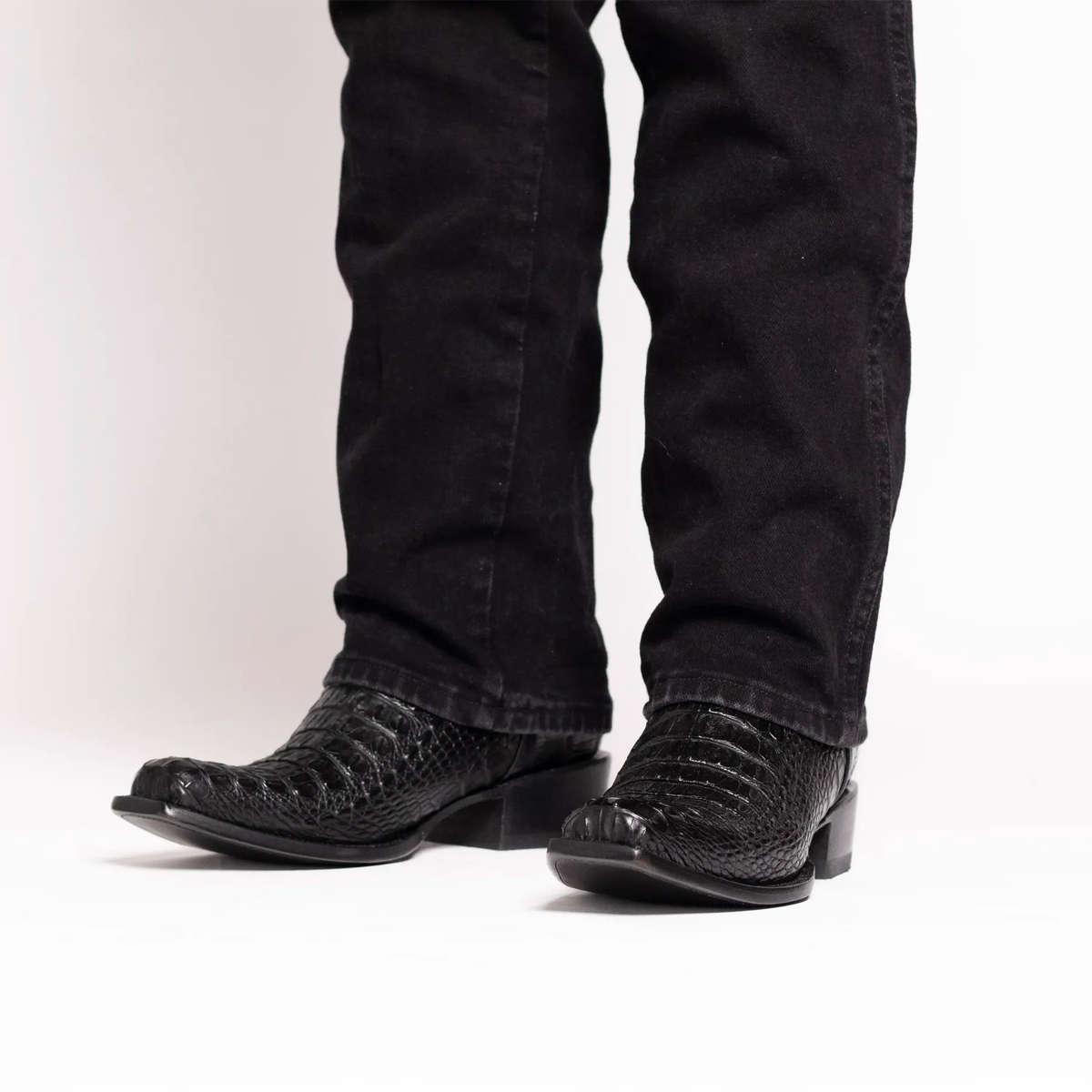 Black alligator cowboy boots worn with black jeans, shown from the knees down on a white studio background.